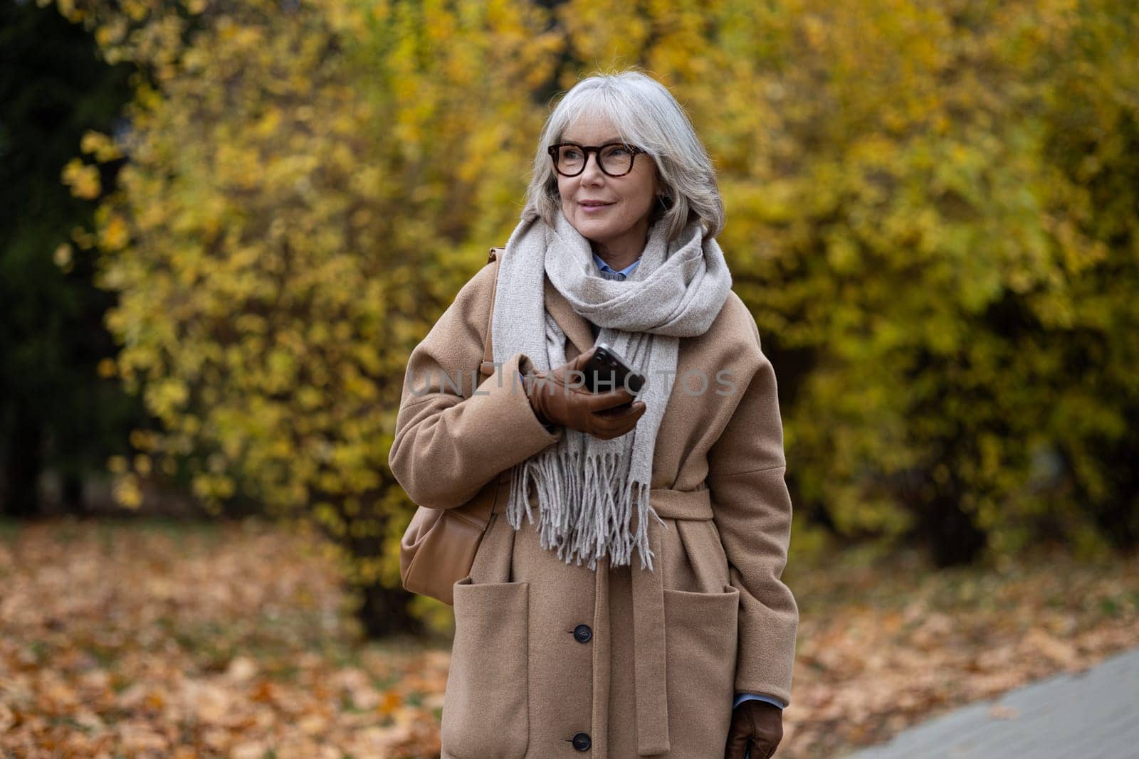 The elderly woman walks casually through a park filled with colorful autumn foliage. She is dressed warmly in a coat and scarf, holding her phone as she smiles.
