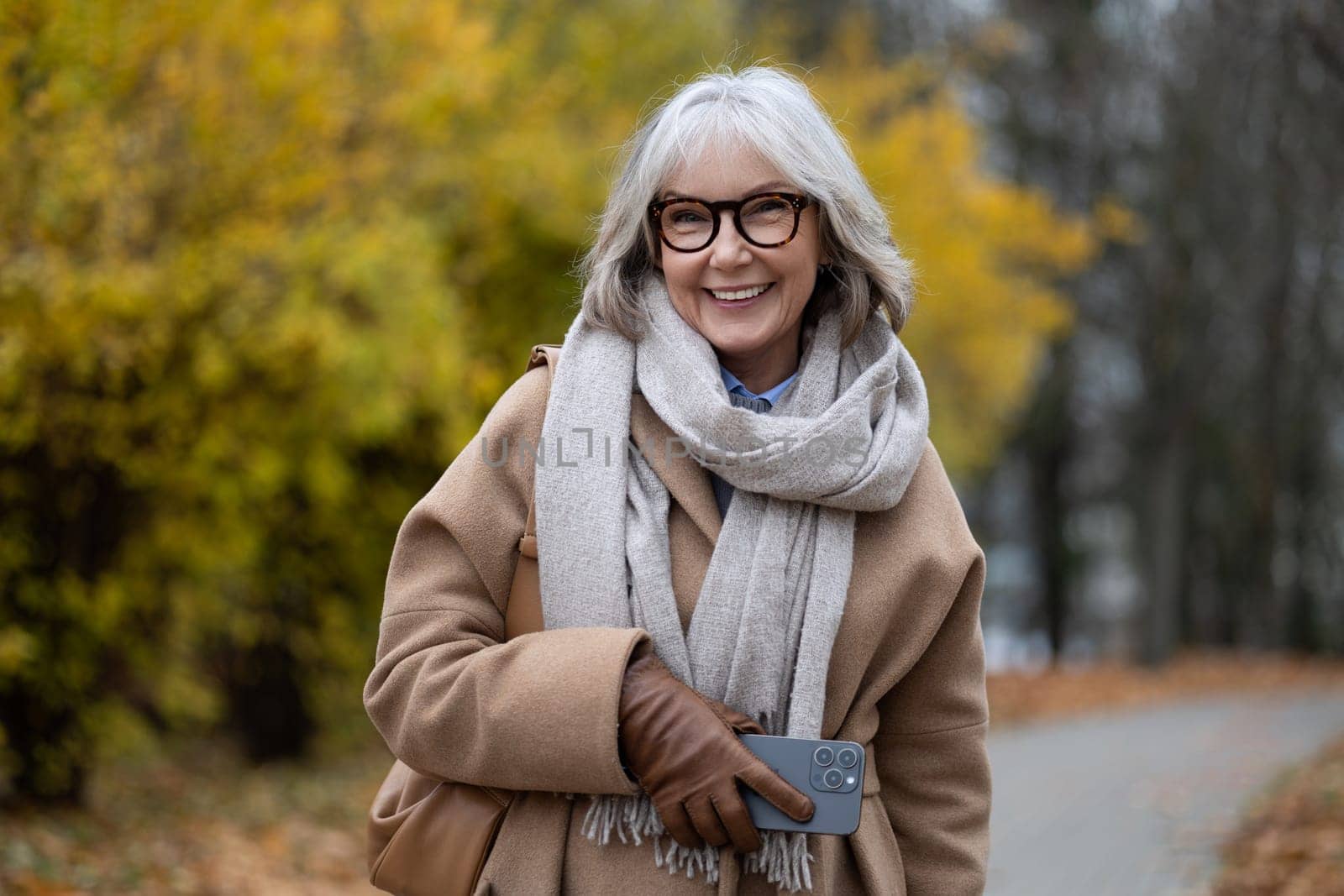An older woman with gray hair and glasses strolls through a beautiful autumn park. She is smiling, wearing a cozy coat and scarf, holding a smartphone in her hand.