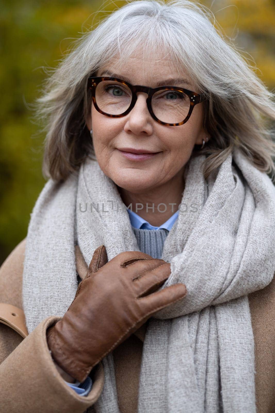 A mature woman stands outdoors on a crisp autumn day, wearing a cozy scarf and stylish gloves. Her silver hair and glasses complement her warm smile as she enjoys the fresh air.