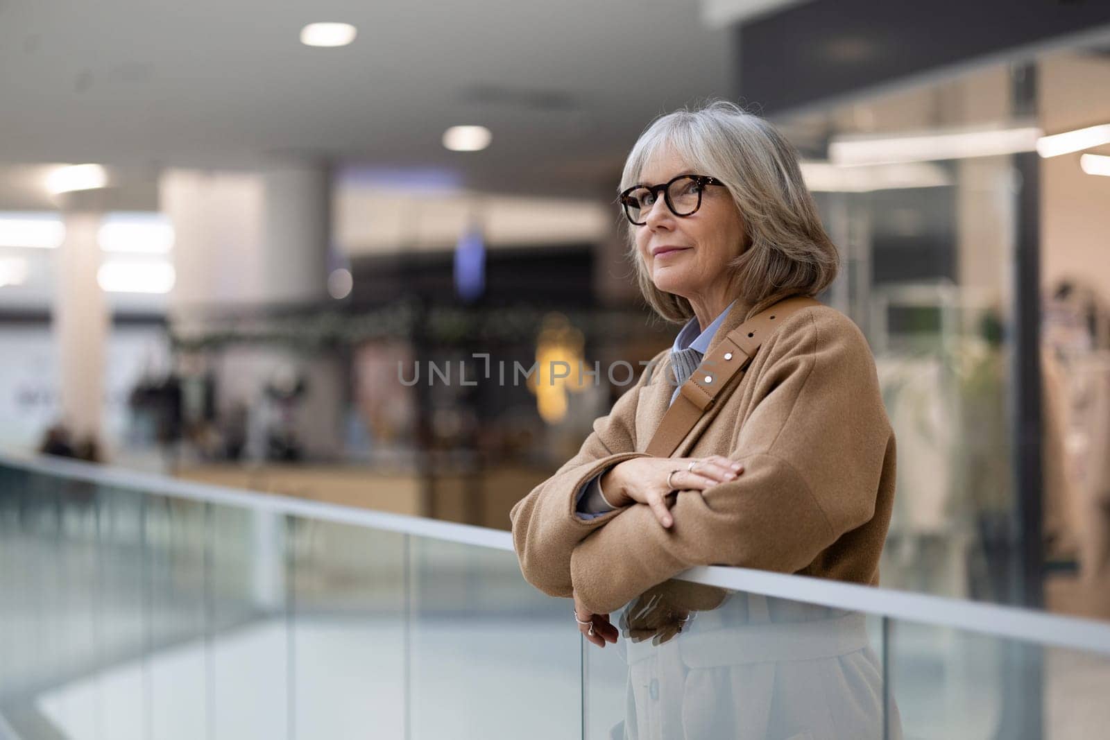 A woman with silver hair and glasses leans against a railing in a shopping mall, appearing contemplative as she looks around, surrounded by modern shops and people.