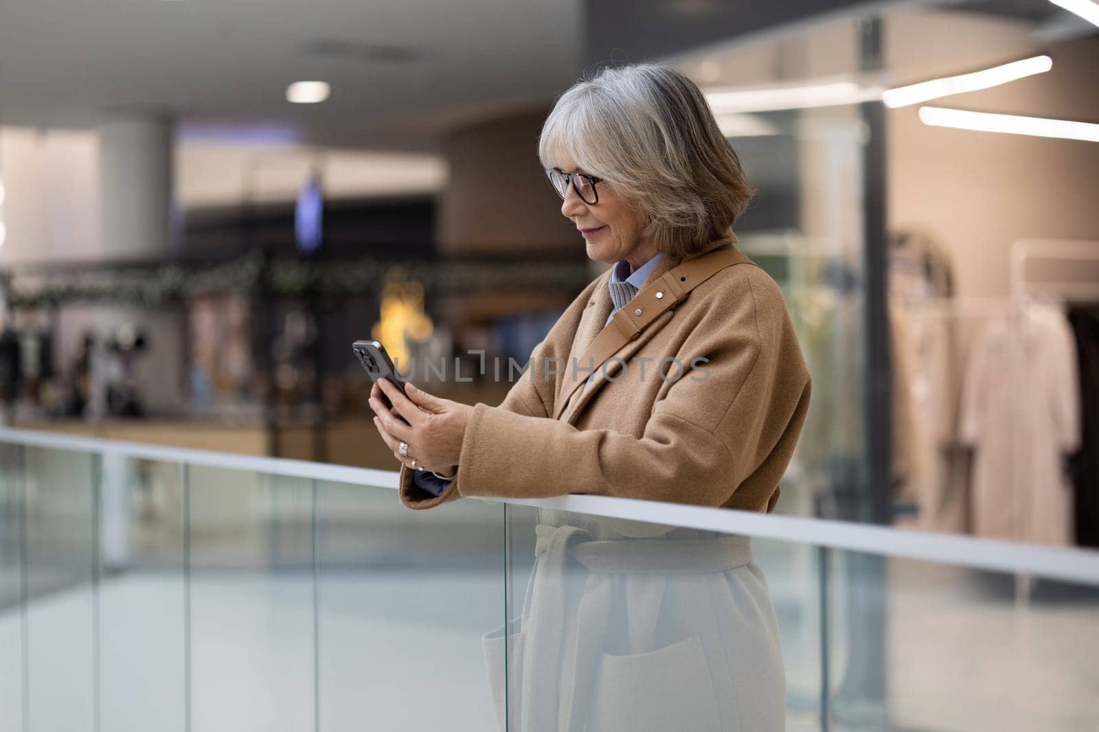 An older woman in a beige coat enjoys scrolling through her smartphone while leaning on a glass railing in a contemporary shopping mall. The atmosphere is bright and inviting.