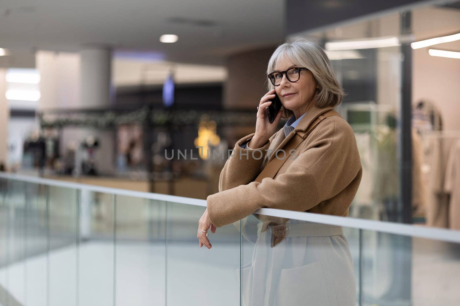 In a bustling shopping mall, a stylish mature woman with glasses engages in a phone conversation while casually leaning on a railing, showcasing a contemporary urban atmosphere.