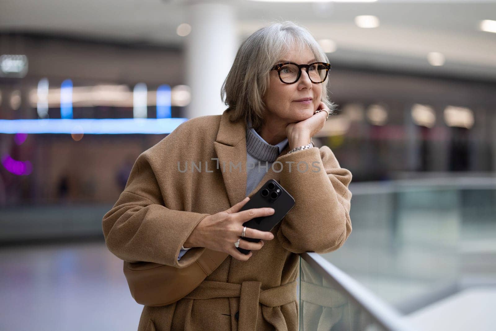 A woman with stylish glasses leans against a railing, thoughtfully holding her smartphone. She appears lost in contemplation in a contemporary setting filled with soft lighting.