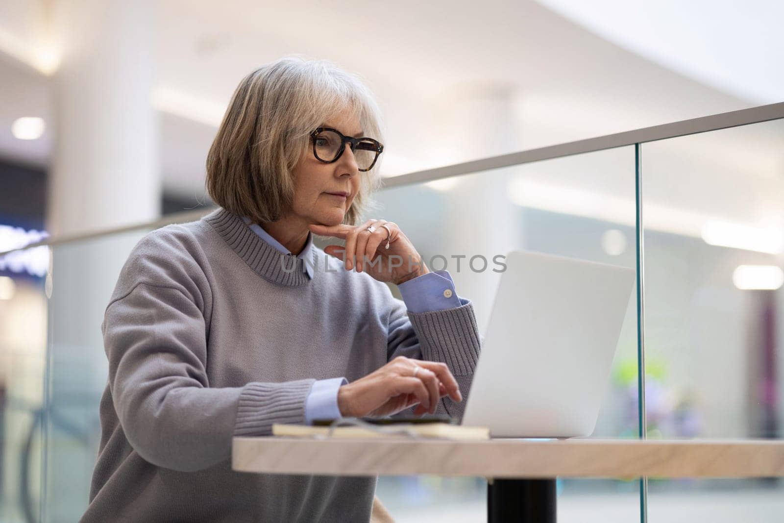 A senior woman sits comfortably at a table in a modern shopping mall cafe, typing on her laptop. She appears focused and engaged in her work while enjoying the atmosphere.