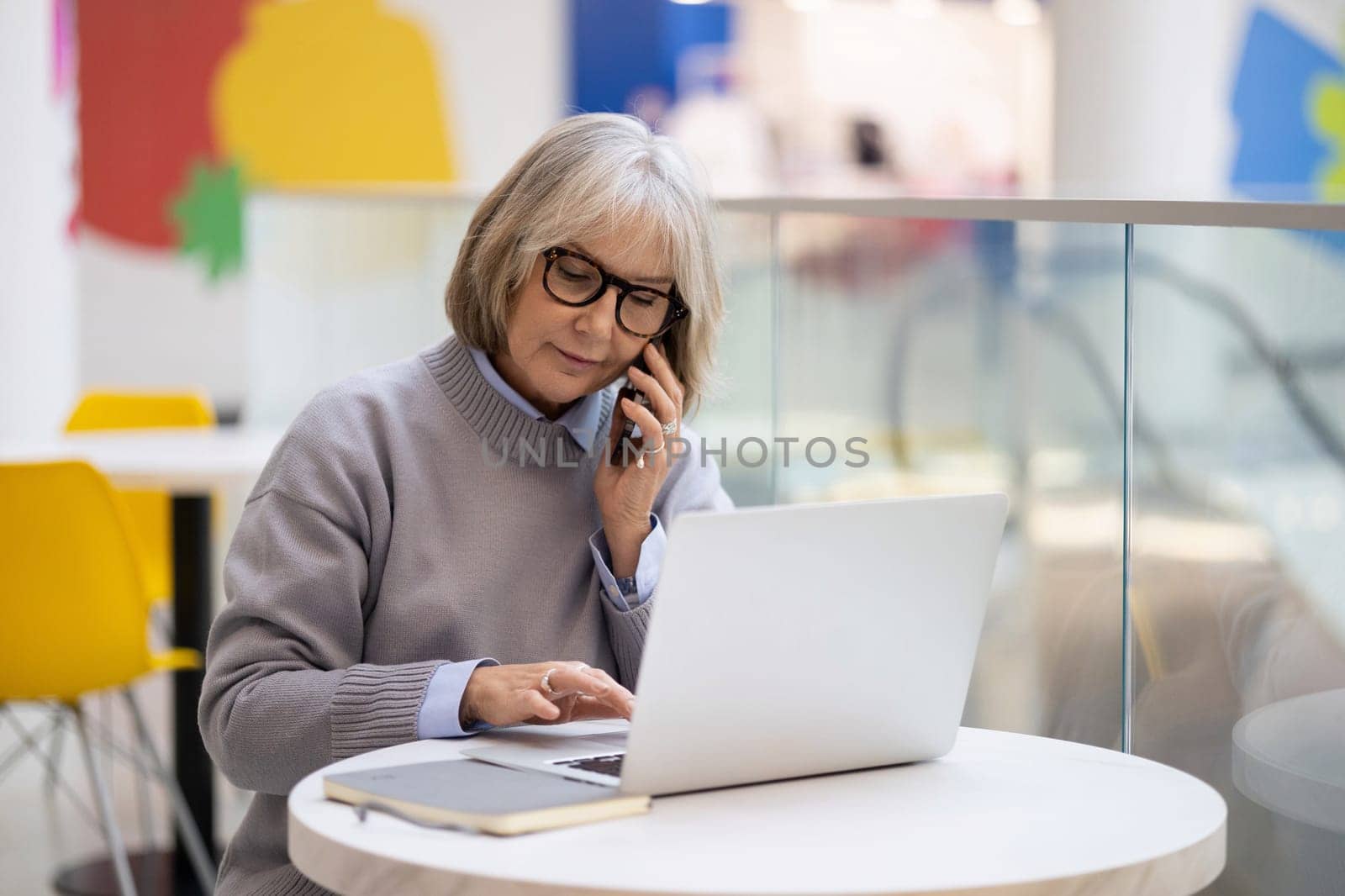 An older woman with glasses sits at a round table, using her laptop while engaged in a phone call. The bright and colorful cafe interior adds a lively atmosphere.