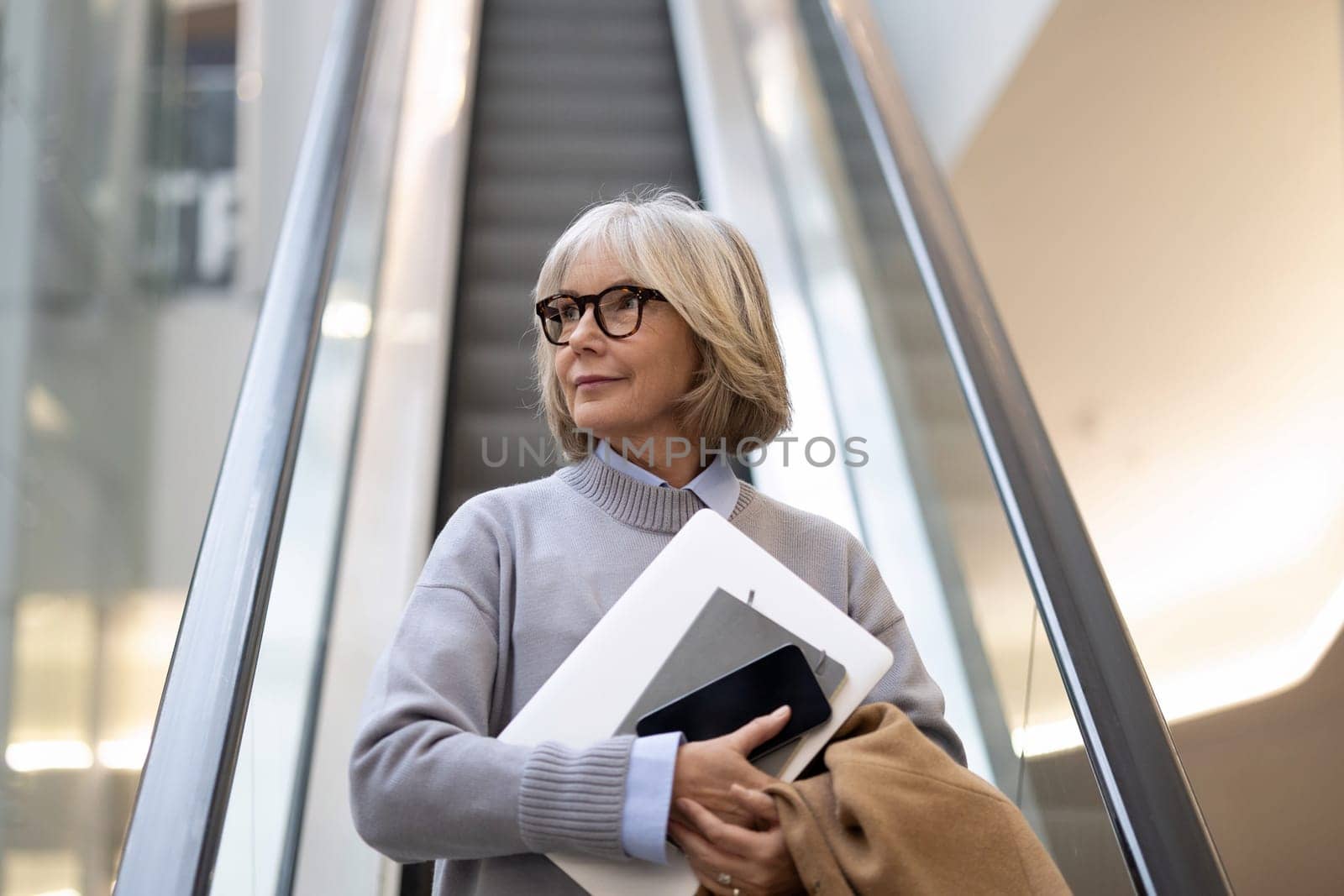 A senior woman with short gray hair is riding an escalator in a contemporary building. She holds a laptop and a notebook, dressed in a cozy sweater, appearing thoughtful.