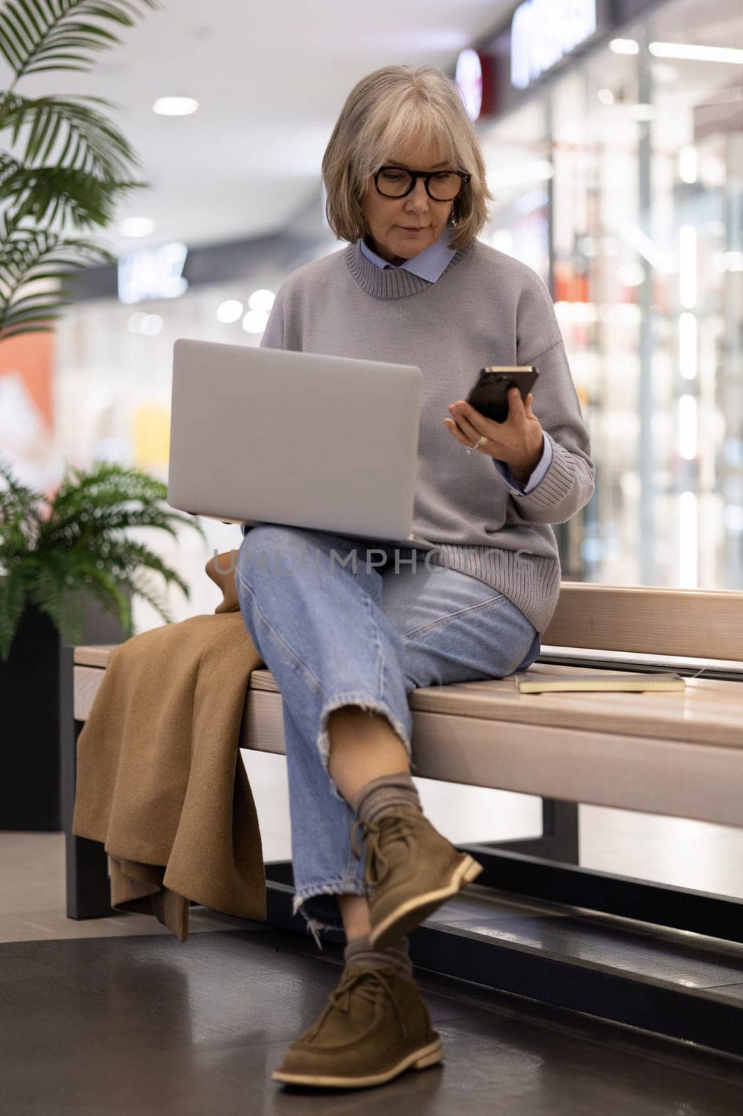 An older woman with glasses sits on a bench in a shopping mall, focused on her laptop and occasionally checking her smartphone, blending leisure with productivity.