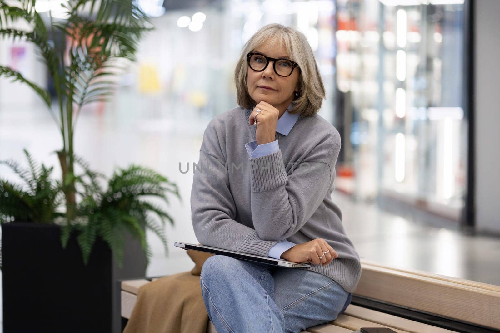 An older woman in a cozy gray sweater sits on a bench in a shopping mall, gazing thoughtfully while holding a notebook, surrounded by lush indoor plants, enjoying her day out.