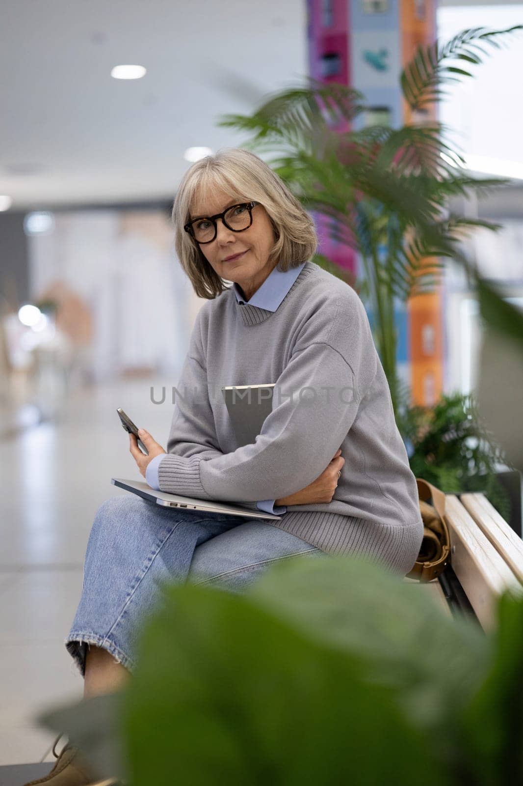 A woman with gray hair and glasses relaxes on a wooden bench, holding a notebook and pen while deep in thought, surrounded by indoor plants and a vibrant backdrop.