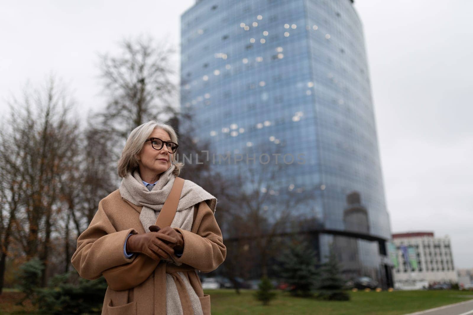 An elderly woman with stylish glasses smiles while standing outside in a brown coat and scarf. The backdrop features a contemporary glass building and bare trees under an overcast sky.