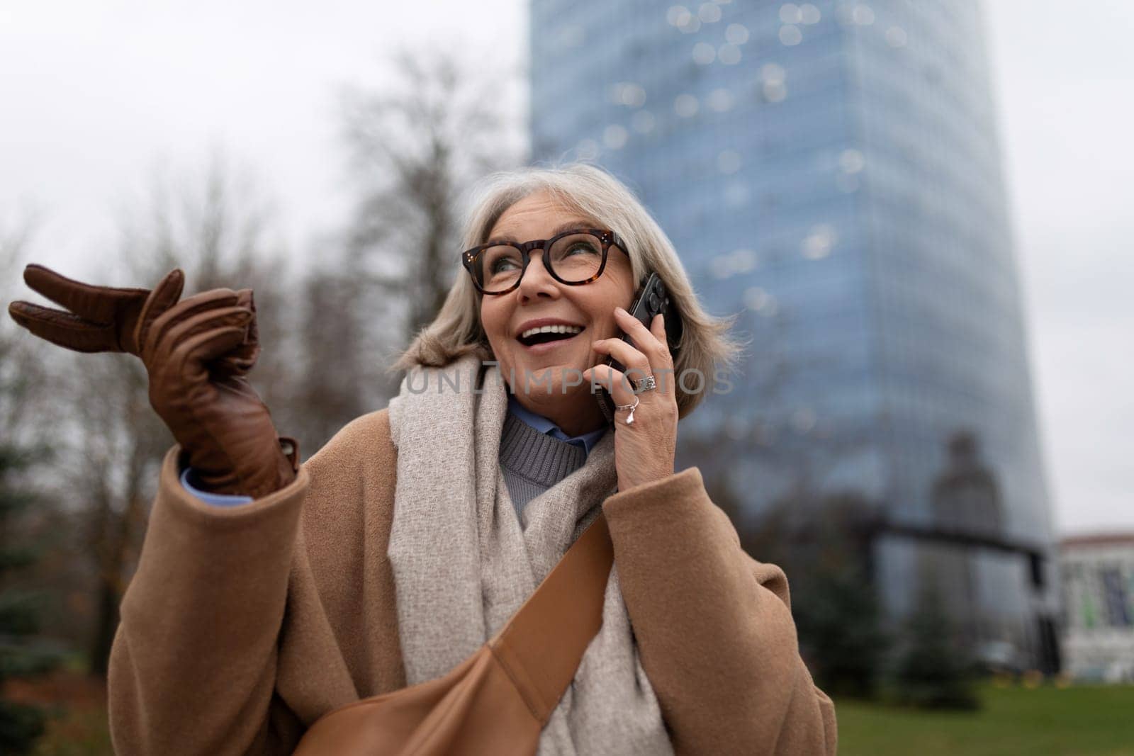 An older woman smiles brightly as she talks on her phone, gesturing joyfully with her hand while standing in a city park. The buildings loom in the background.