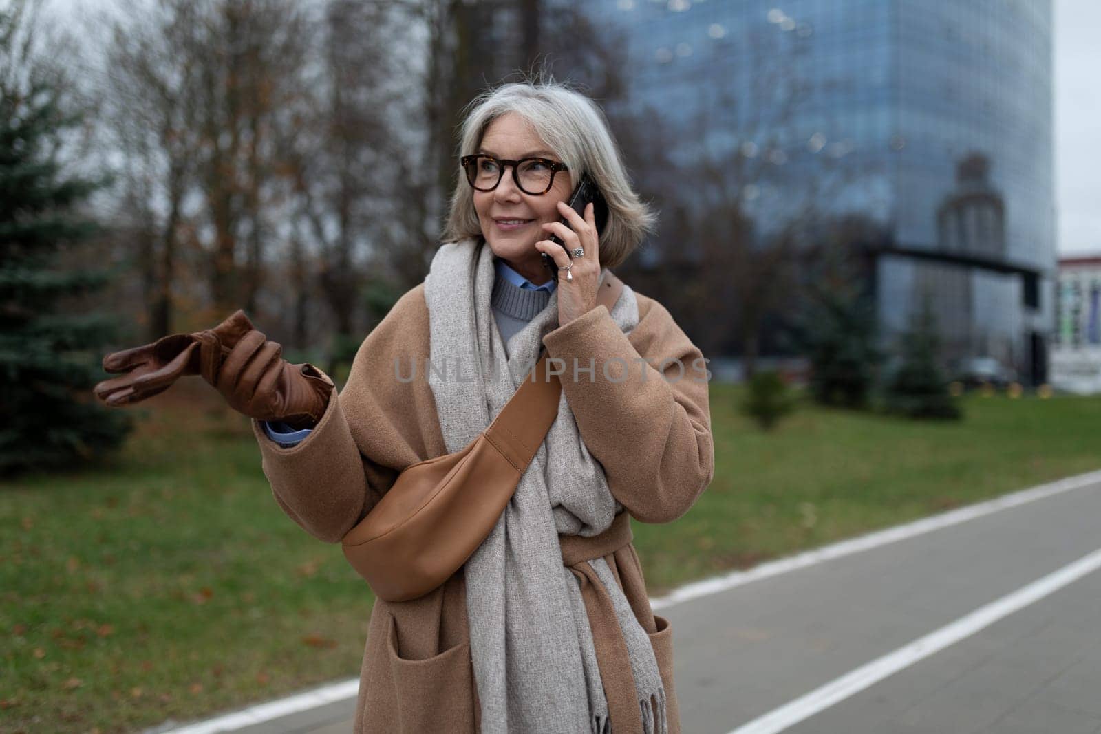 An older woman enjoys a stroll in the park, wrapped in a warm coat and scarf. She chats on her smartphone while holding a pair of gloves, surrounded by autumn scenery.