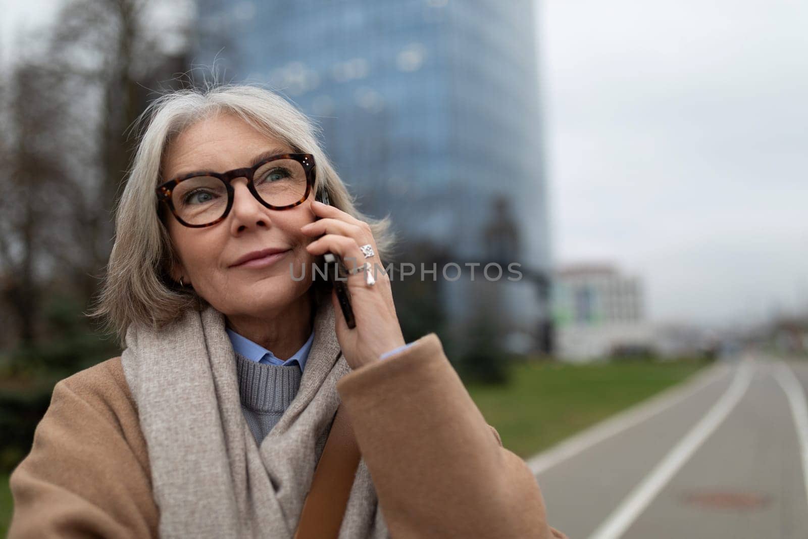 A woman with gray hair enjoys a phone conversation during a stroll in a city park. The overcast sky creates a calm atmosphere as she smiles thoughtfully.