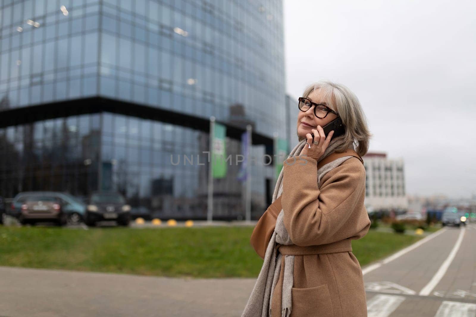 An older woman in a beige coat is engaged in a phone call, looking up thoughtfully while standing near a large glass office building. The weather is overcast and urban.