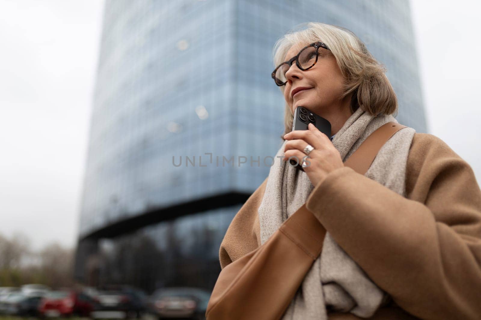 A thoughtful woman dressed warmly is holding a phone near her chin as she gazes upward, surrounded by a modern glass building under a cloudy sky.