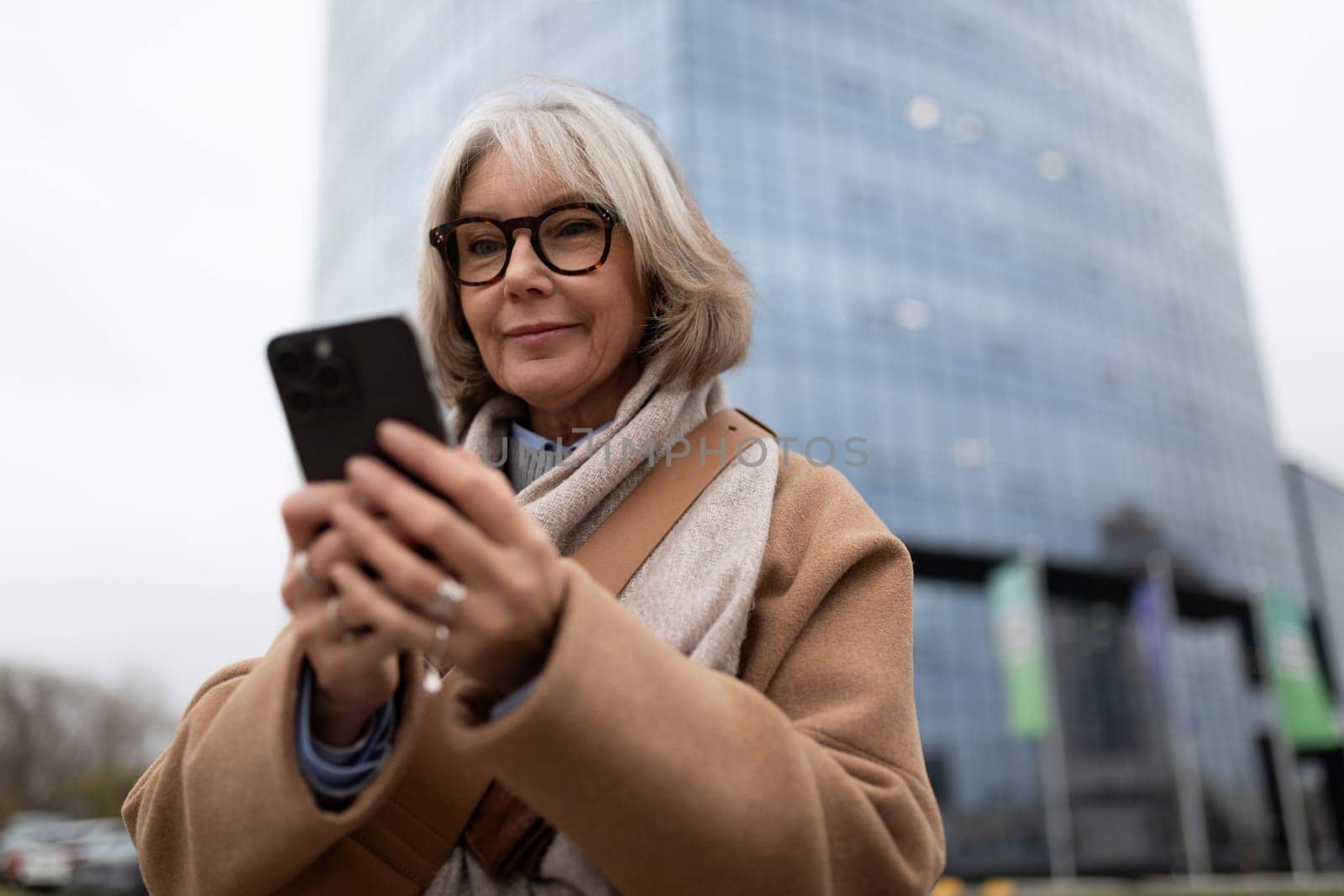 A woman with gray hair and glasses checks her smartphone while standing in front of a sleek, tall building. The weather is overcast, creating a calm atmosphere.