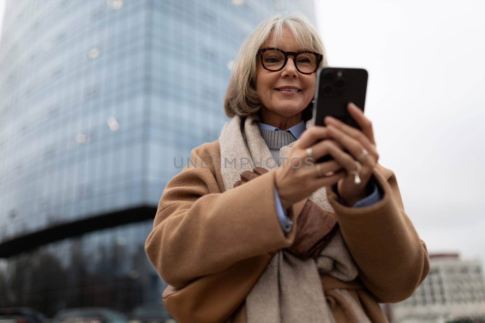 A cheerful older woman with glasses interacts with her smartphone in a city environment. She is dressed warmly, displaying a sense of contentment amidst the urban landscape.