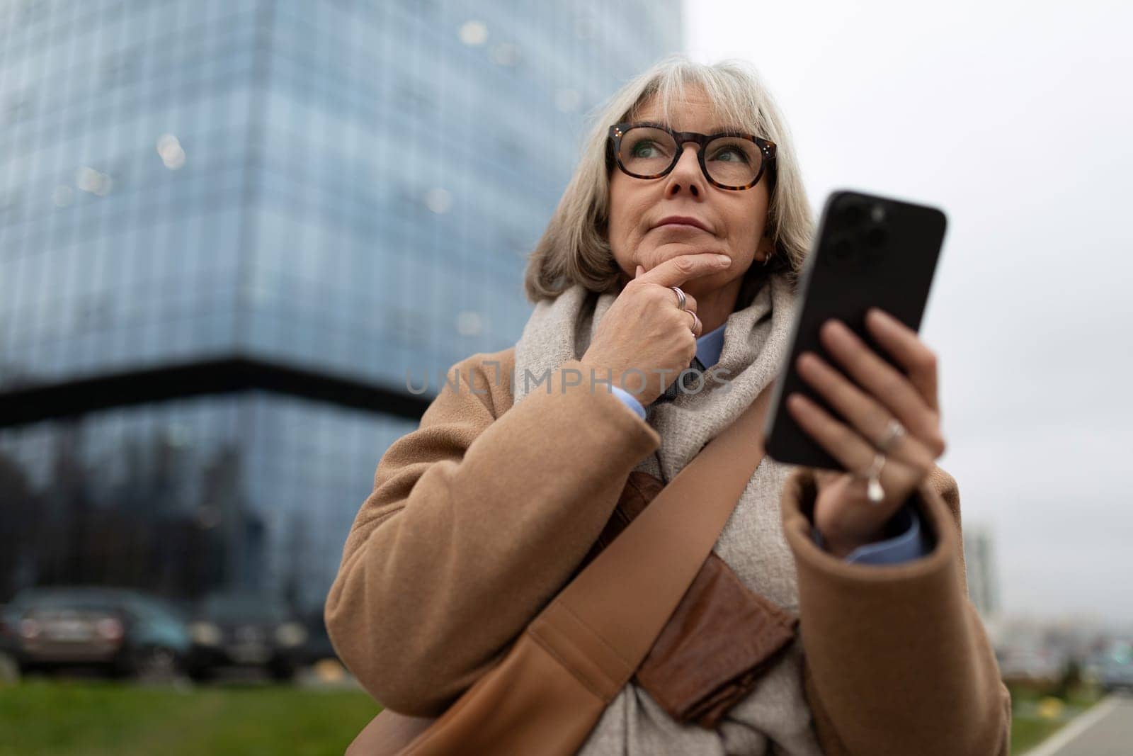 A senior woman stands outdoors, thoughtfully holding her smartphone. She appears to be considering something important, surrounded by a modern urban setting.