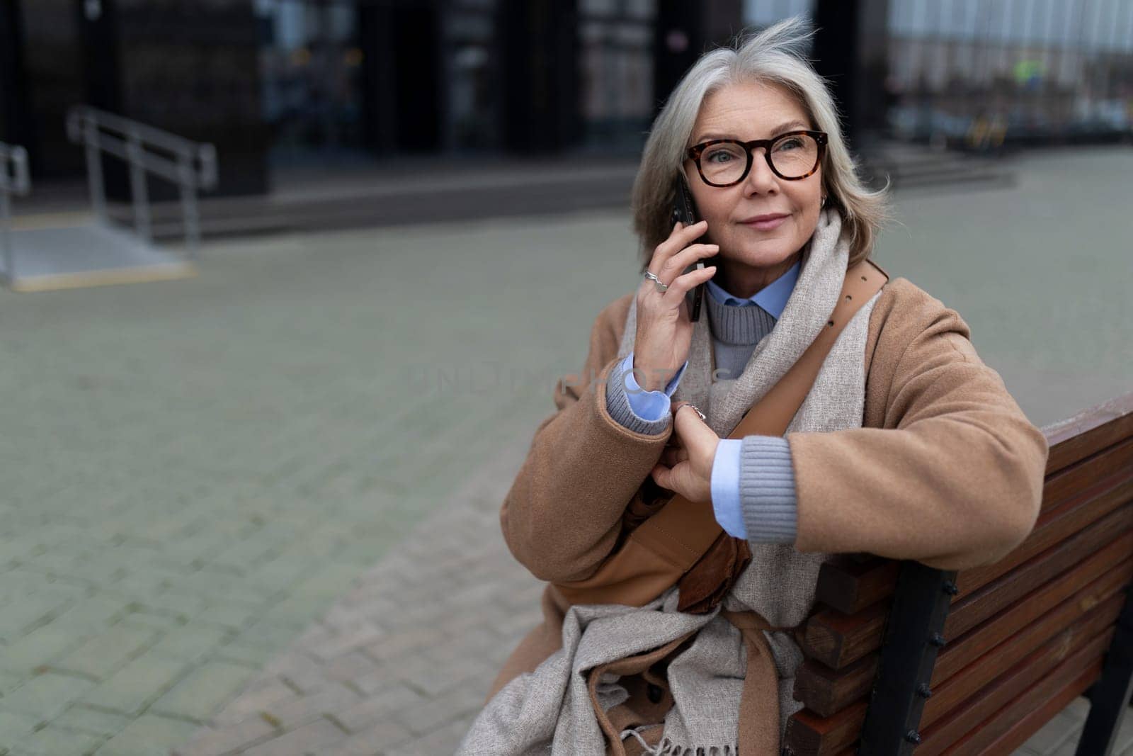 An older woman with gray hair and glasses sits relaxed on a bench, talking on her phone. She is dressed warmly for the cool weather in an urban area.