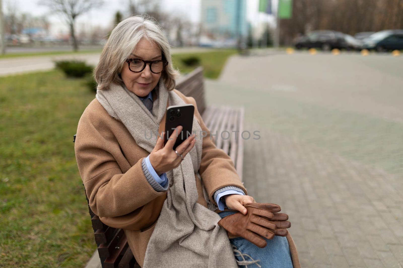 A middle-aged woman with gray hair and glasses dressed in a warm coat and scarf sits comfortably on a park bench. She is focused on her phone, enjoying a chilly afternoon.