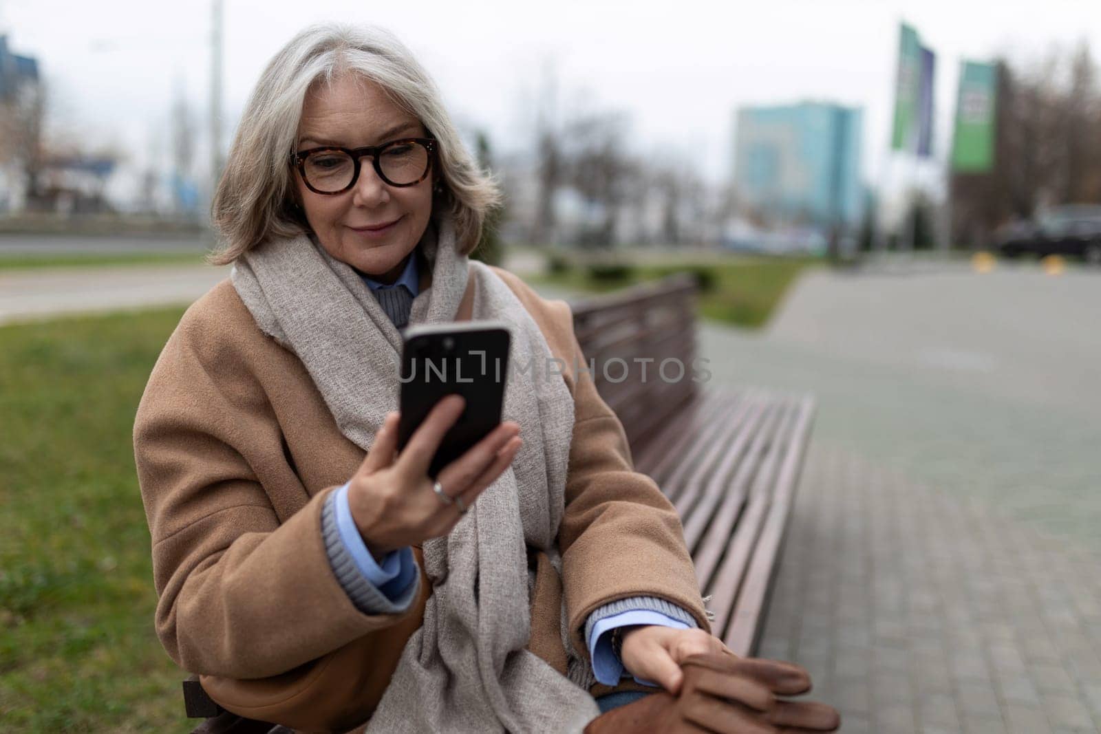 A woman with gray hair and glasses sits comfortably on a park bench, smiling as she interacts with her smartphone in a city environment during springtime.