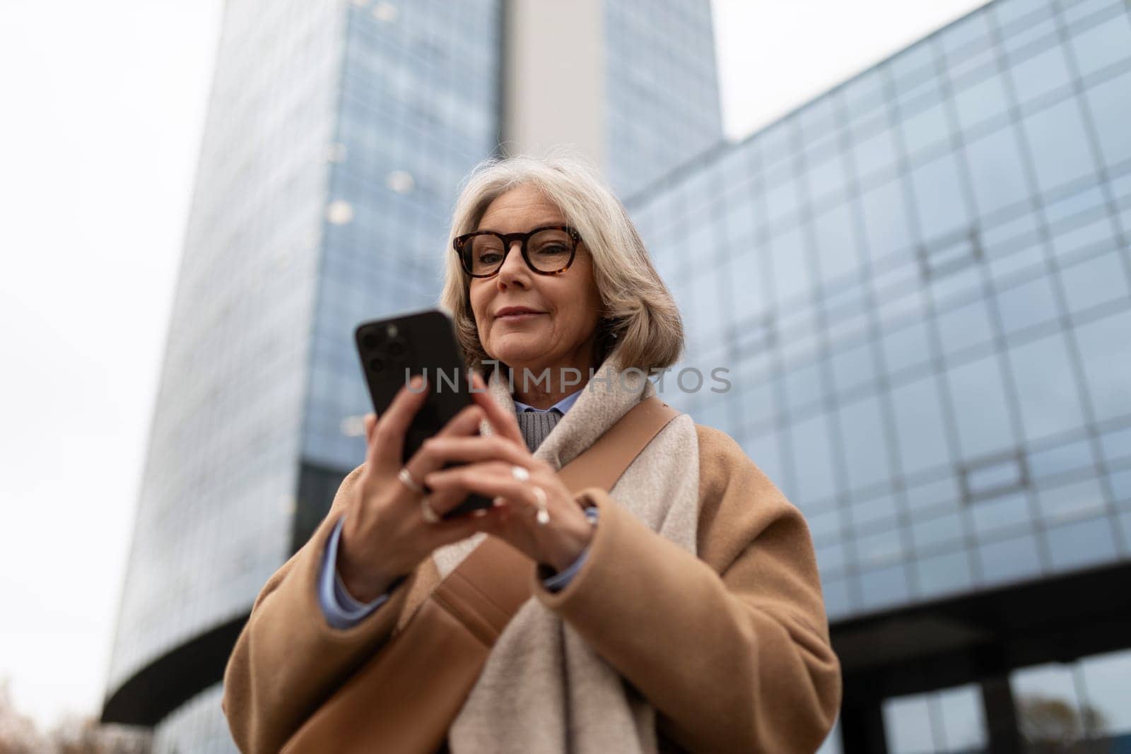 A woman with gray hair, wearing glasses and a stylish coat, focuses on her smartphone amidst urban architecture on an overcast day.
