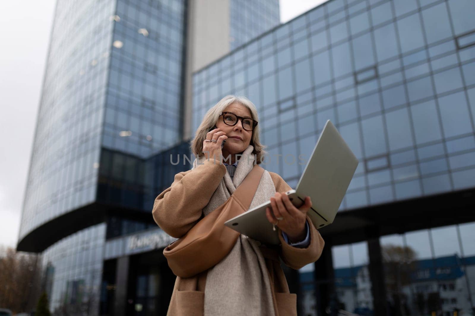 A businesswoman stands in front of a modern glass office building, engaged in a phone conversation while working on her laptop. The day is cloudy, hinting at a busy work environment.