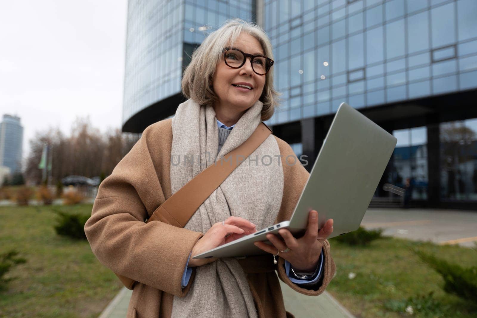A senior woman stands outside a contemporary office building, smiling as she types on her laptop. The setting reflects a blend of nature and urban life on a cool day.
