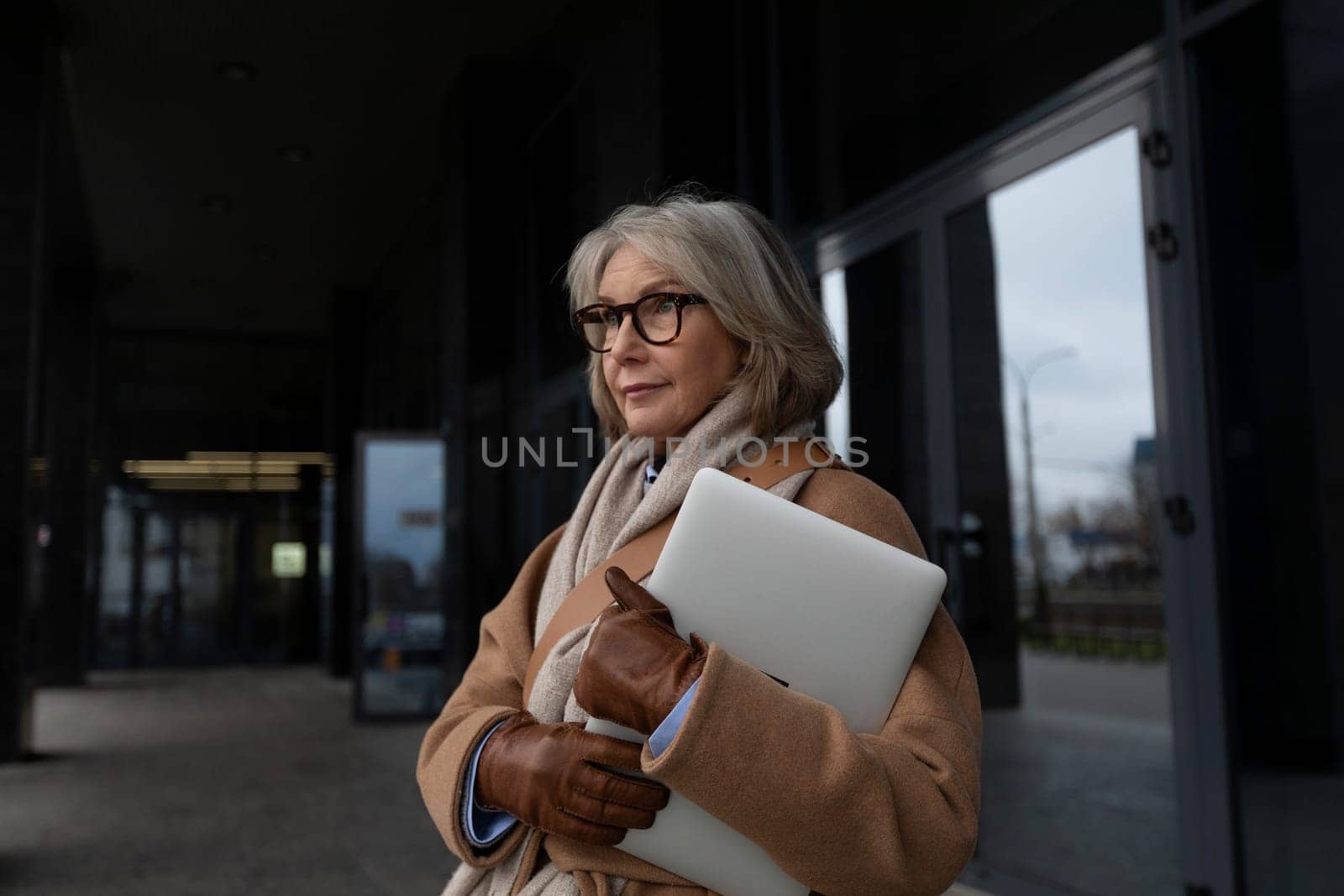 A stylish woman with gray hair stands outside a contemporary building. She holds a laptop while dressed warmly in a coat and scarf, reflecting a thoughtful expression.