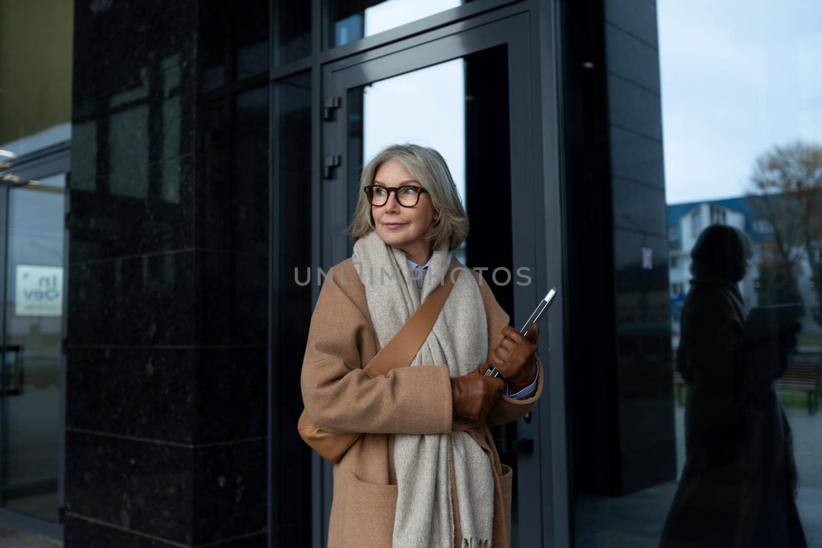 An older woman with gray hair stands outside a modern building, dressed in a cozy coat and scarf, holding a phone. She gazes thoughtfully into the distance, enjoying a chilly day.