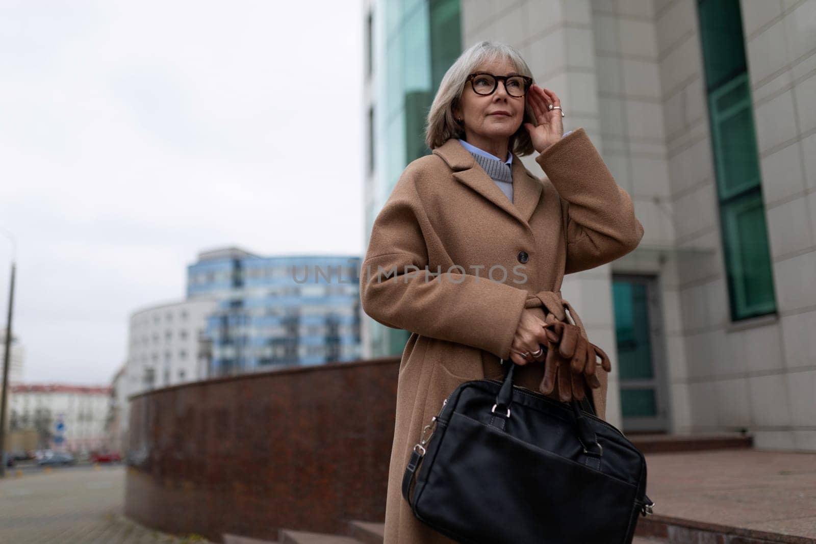 A senior woman dressed in a fashionable coat and glasses holds a handbag while standing near a contemporary building. She appears thoughtful as she adjusts her hair on a cloudy day.