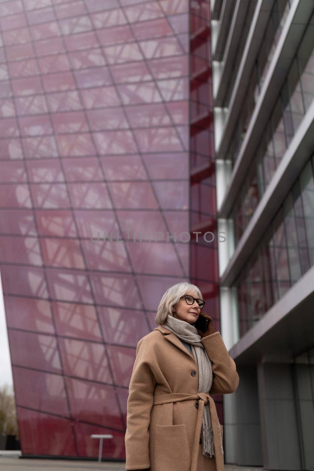 The woman walks confidently while speaking on her phone, dressed in a stylish beige coat and scarf. She stands beside a contemporary building with a geometric, purple exterior.