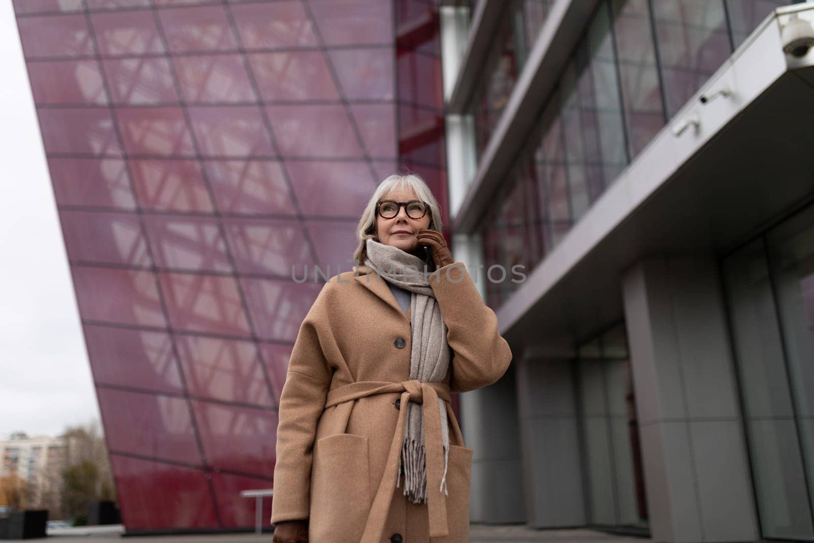 A woman with gray hair wearing glasses stands outdoors, looking thoughtfully while dressed in a cozy coat and scarf. The backdrop features modern architectural elements.