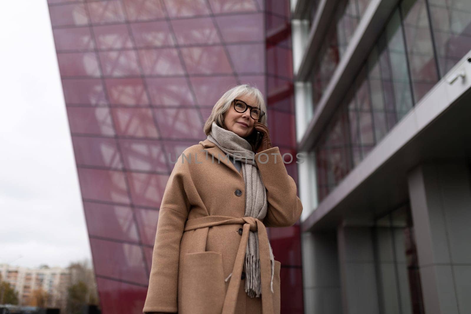 A woman with gray hair and glasses wears a fashionable coat and scarf while standing near a contemporary architectural structure. It is a cool, overcast day.