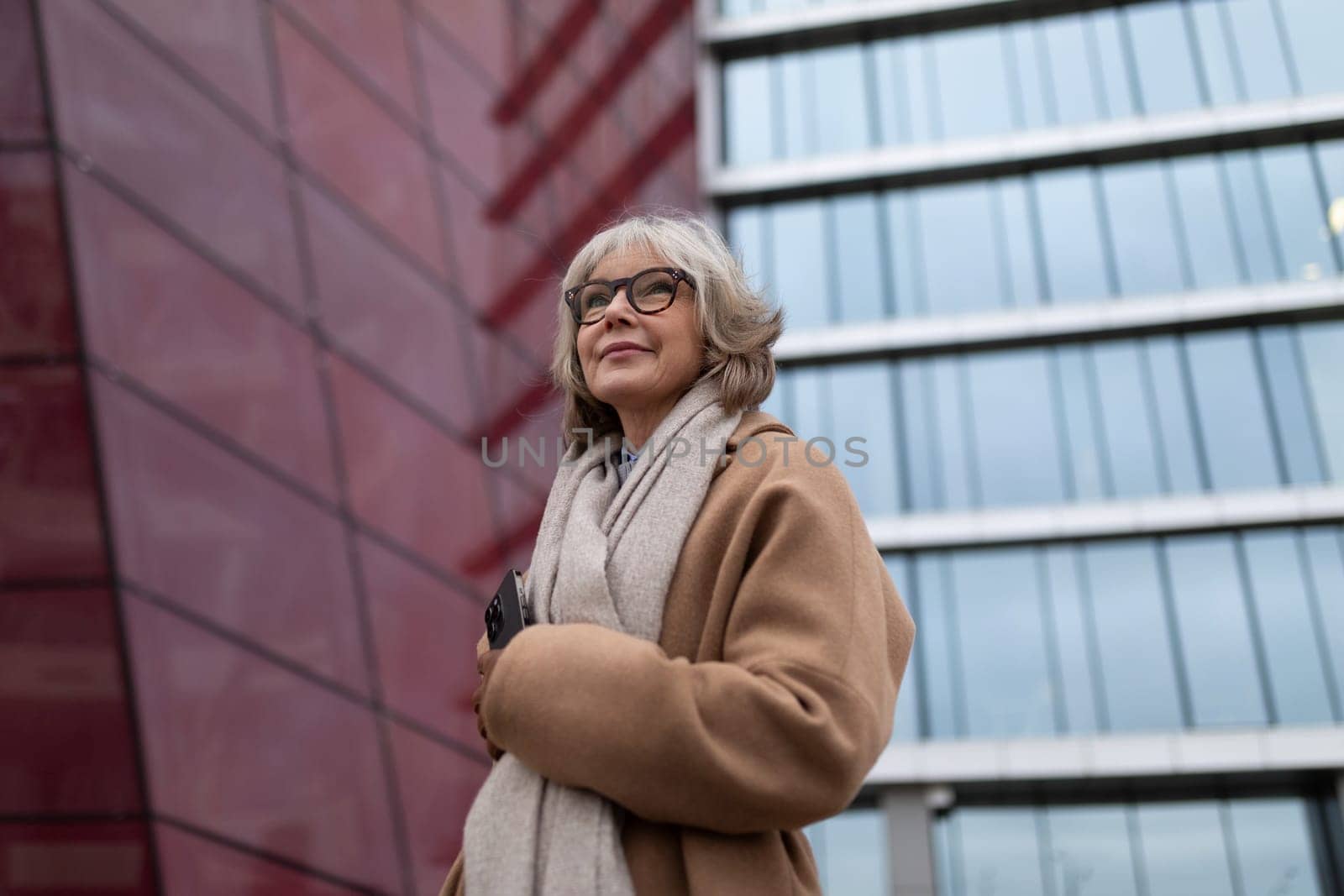 An older woman with glasses and gray hair stands confidently near a large glass building. She wears a cozy coat and holds her phone, enjoying the cool weather.