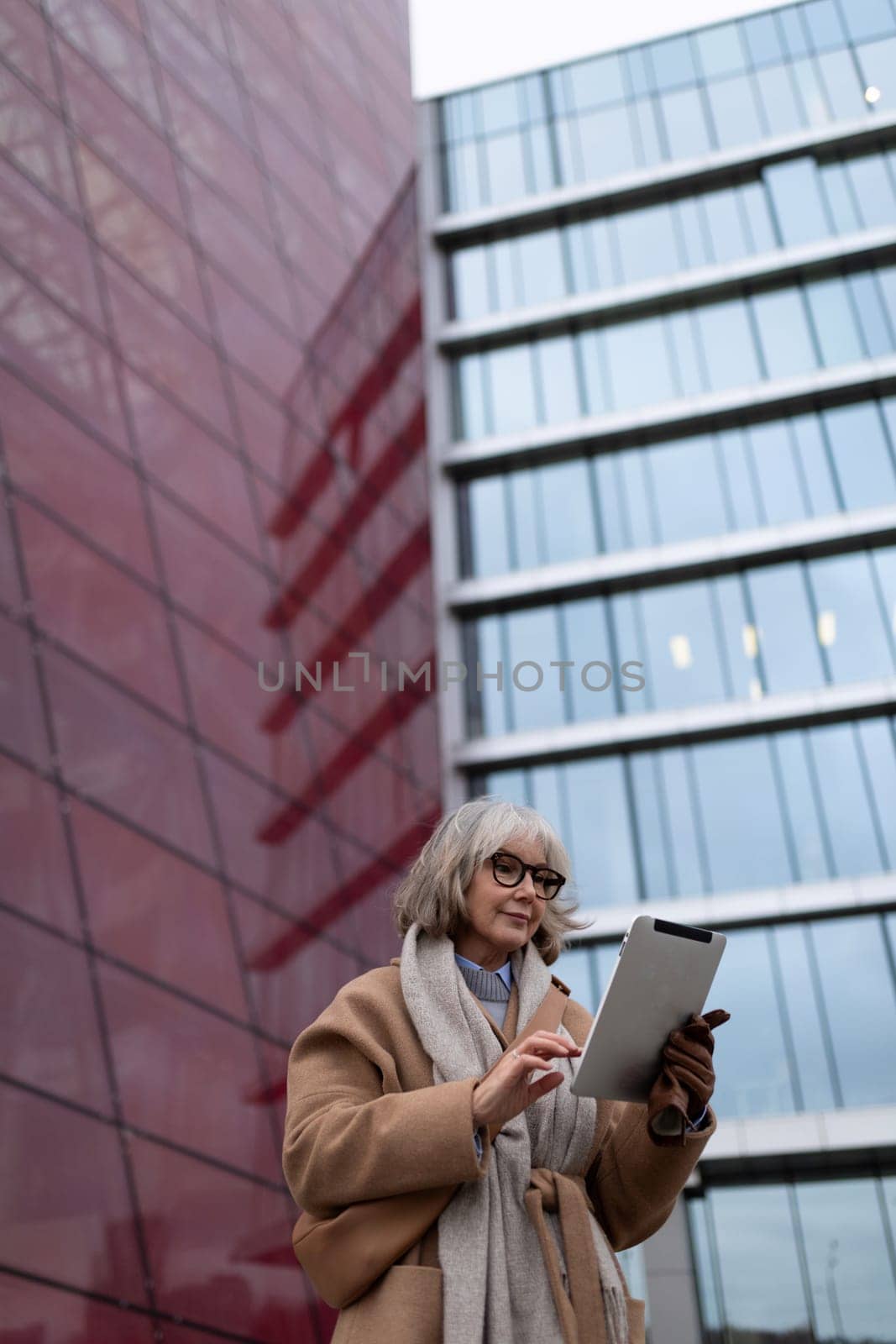A woman with gray hair and glasses stands in an urban setting, focused on her tablet. The striking red and glass architecture surrounds her as she engages with technology.