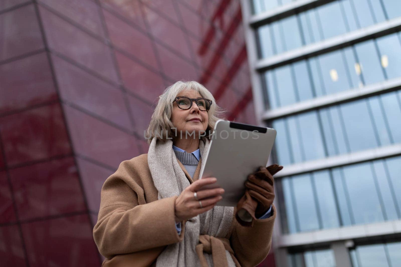 A woman with glasses and gray hair stands outside, holding a tablet. She wears a beige coat and gray scarf, focused on her device amid the striking architecture.