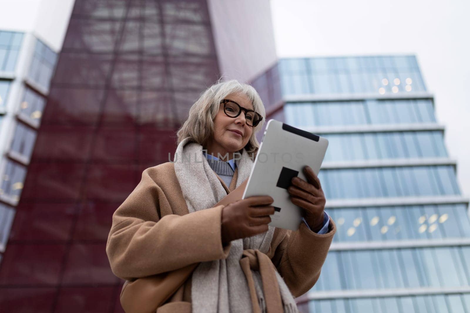 A senior woman is engaged with a tablet, standing outside contemporary buildings. The reflective glass and bold structure create a striking backdrop as twilight descends.