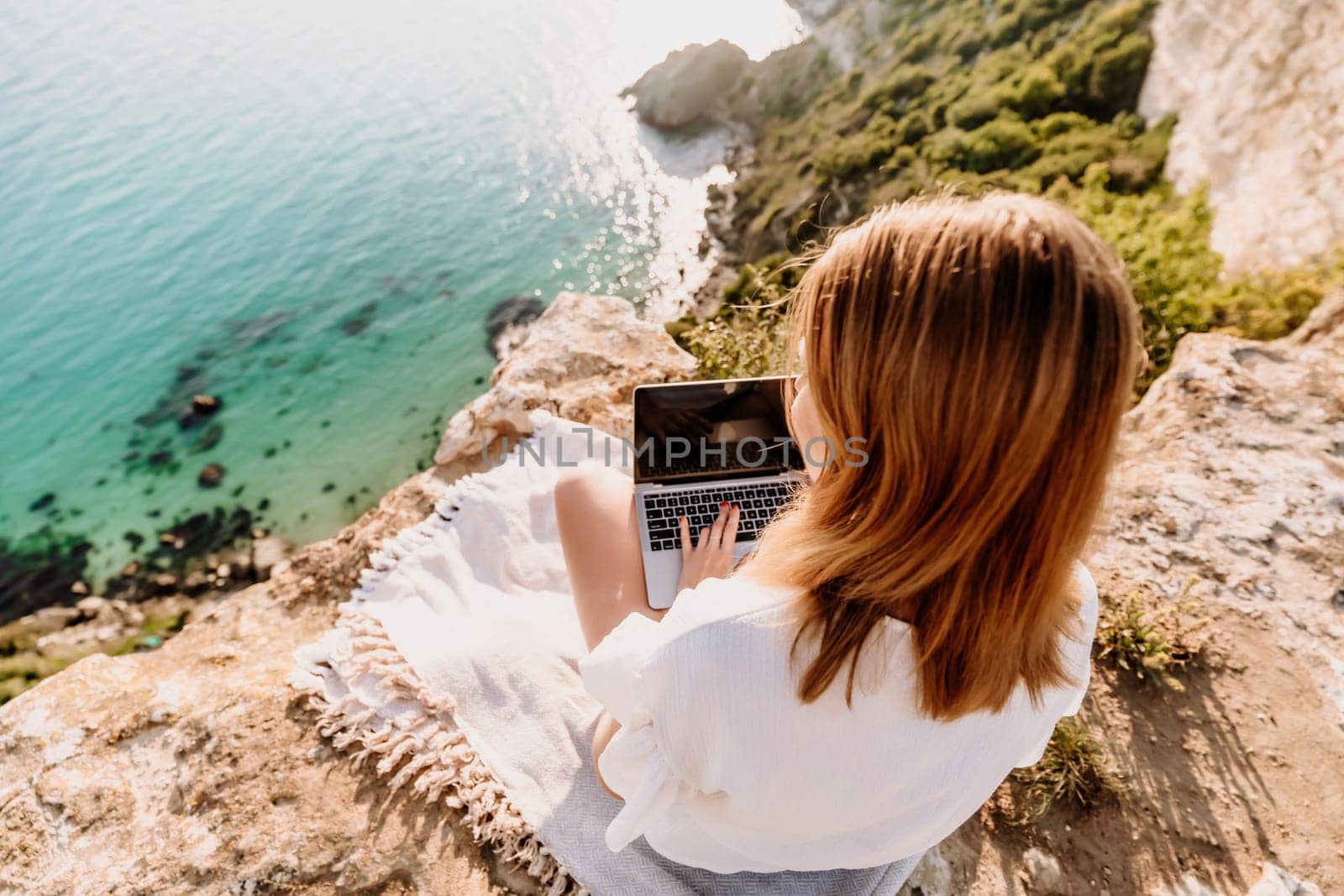 A woman is sitting on a rock overlooking the ocean with a laptop in front of her. She is enjoying the view and the peacefulness of the location