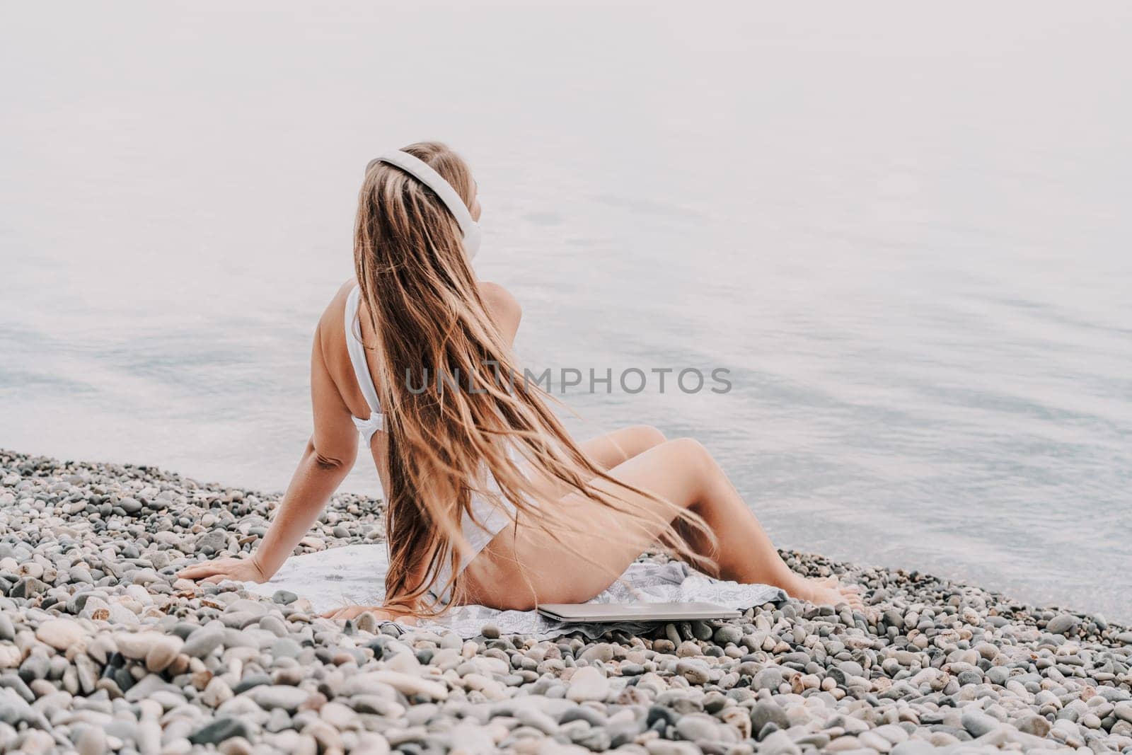 A woman is sitting on a beach with her headphones on. She is wearing a white bikini and a white shirt. The beach is rocky and the water is calm. The woman is enjoying her time by the water