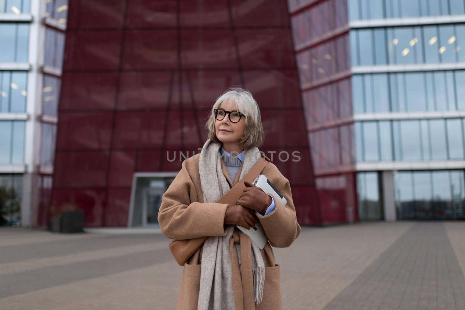 An elegant older woman holds a folder while dressed in a stylish coat and scarf, standing in front of an impressive modern building with expansive glass windows, enjoying a clear day.