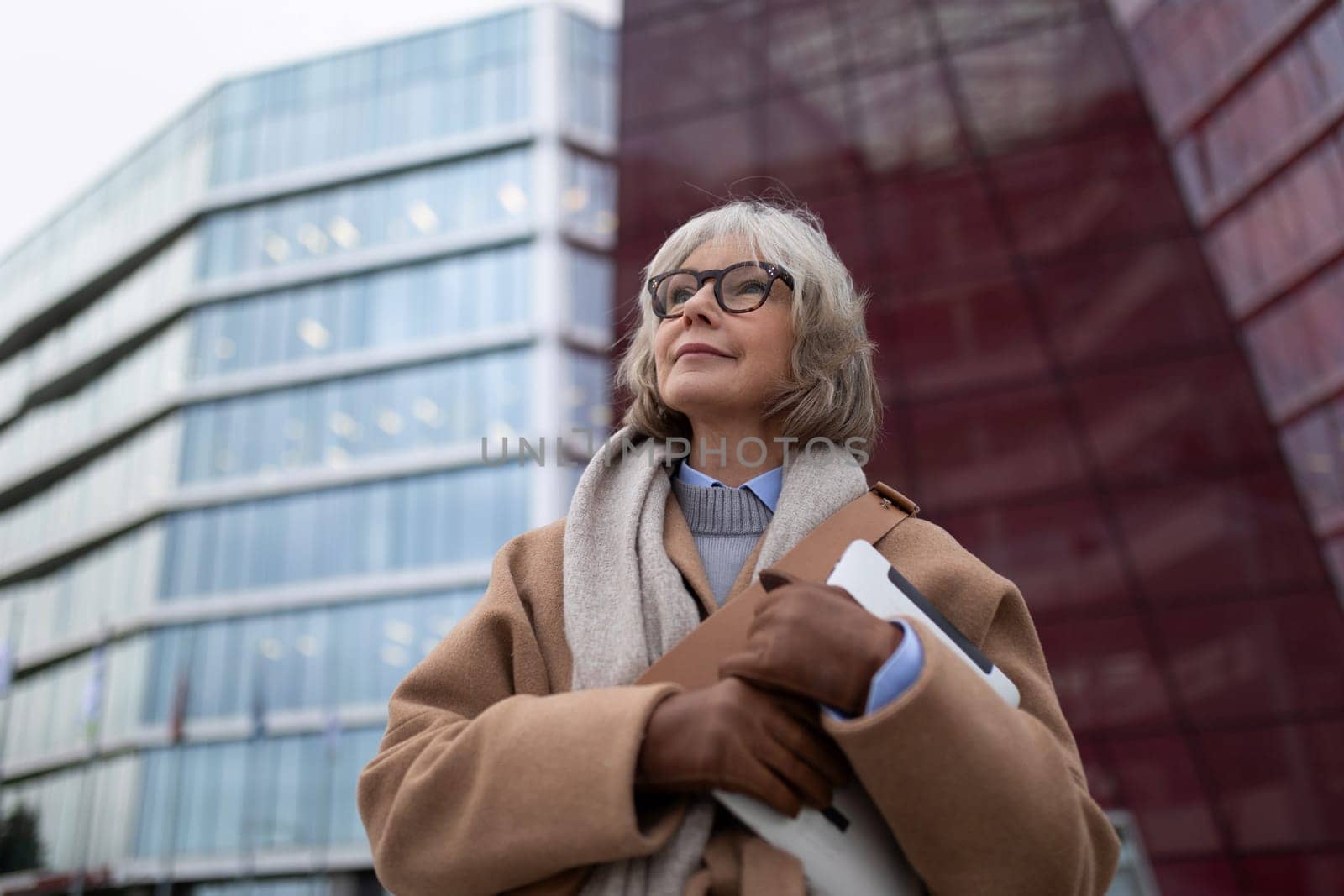 An older woman stands outside modern glass buildings, confidently holding a laptop and wearing a stylish tan coat and scarf. The cloudy sky sets a reflective tone for the setting.