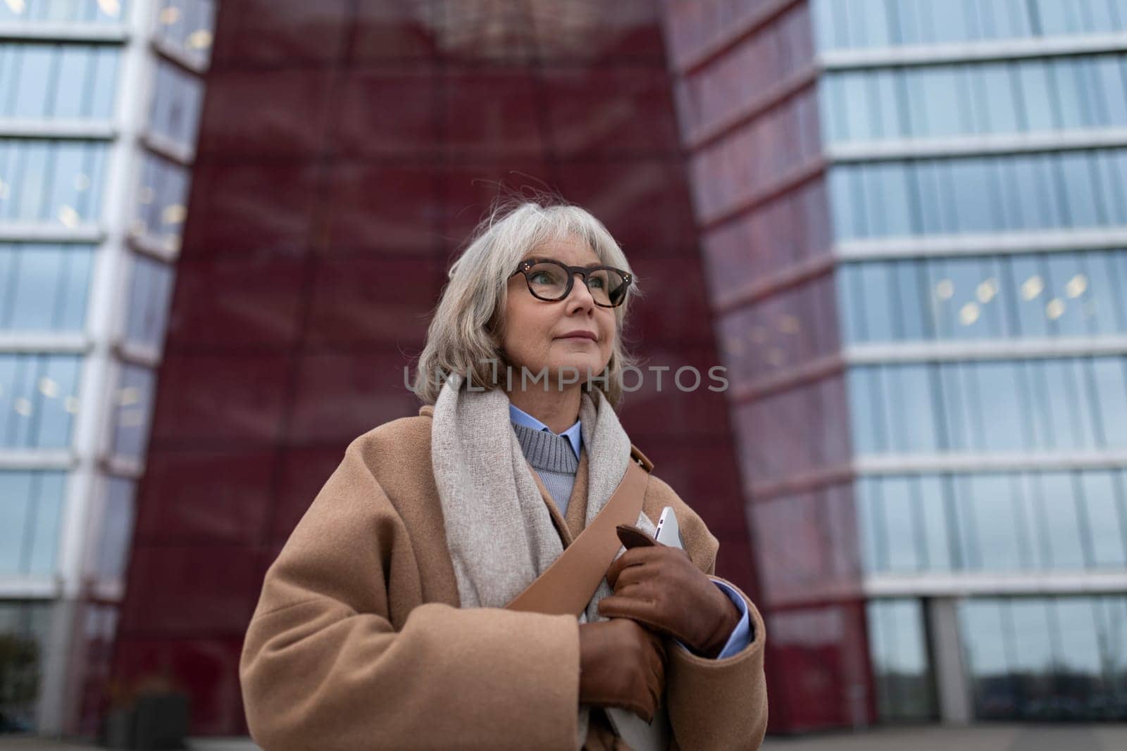 A senior woman with short gray hair and glasses stands outside a striking modern structure. She wears a cozy coat and holds a bag, looking thoughtful and assured amidst the urban setting.