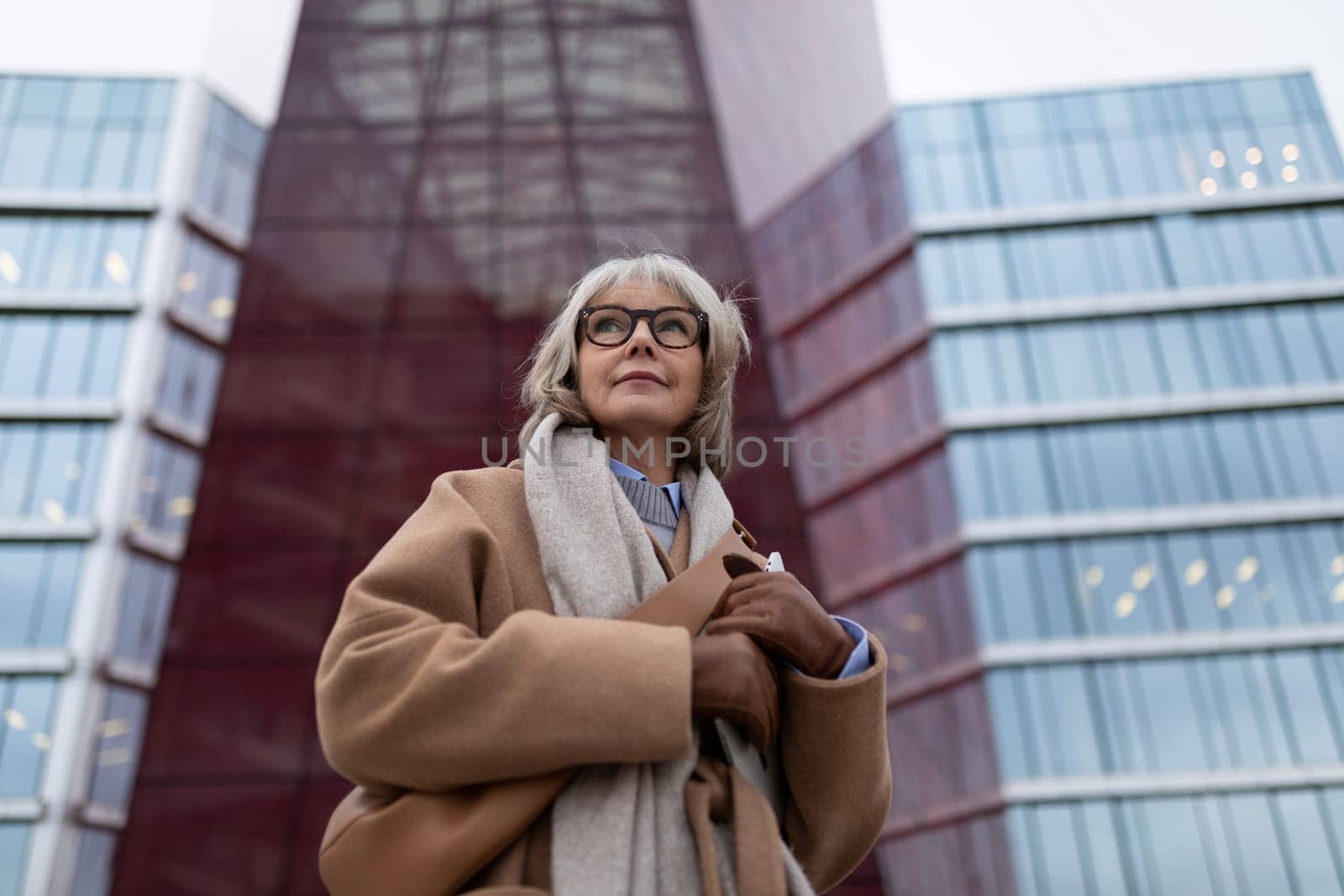 The senior woman stands gracefully with her arms crossed, dressed in a warm coat and scarf, showcasing her elegant style against a contemporary architecture backdrop during dusk.