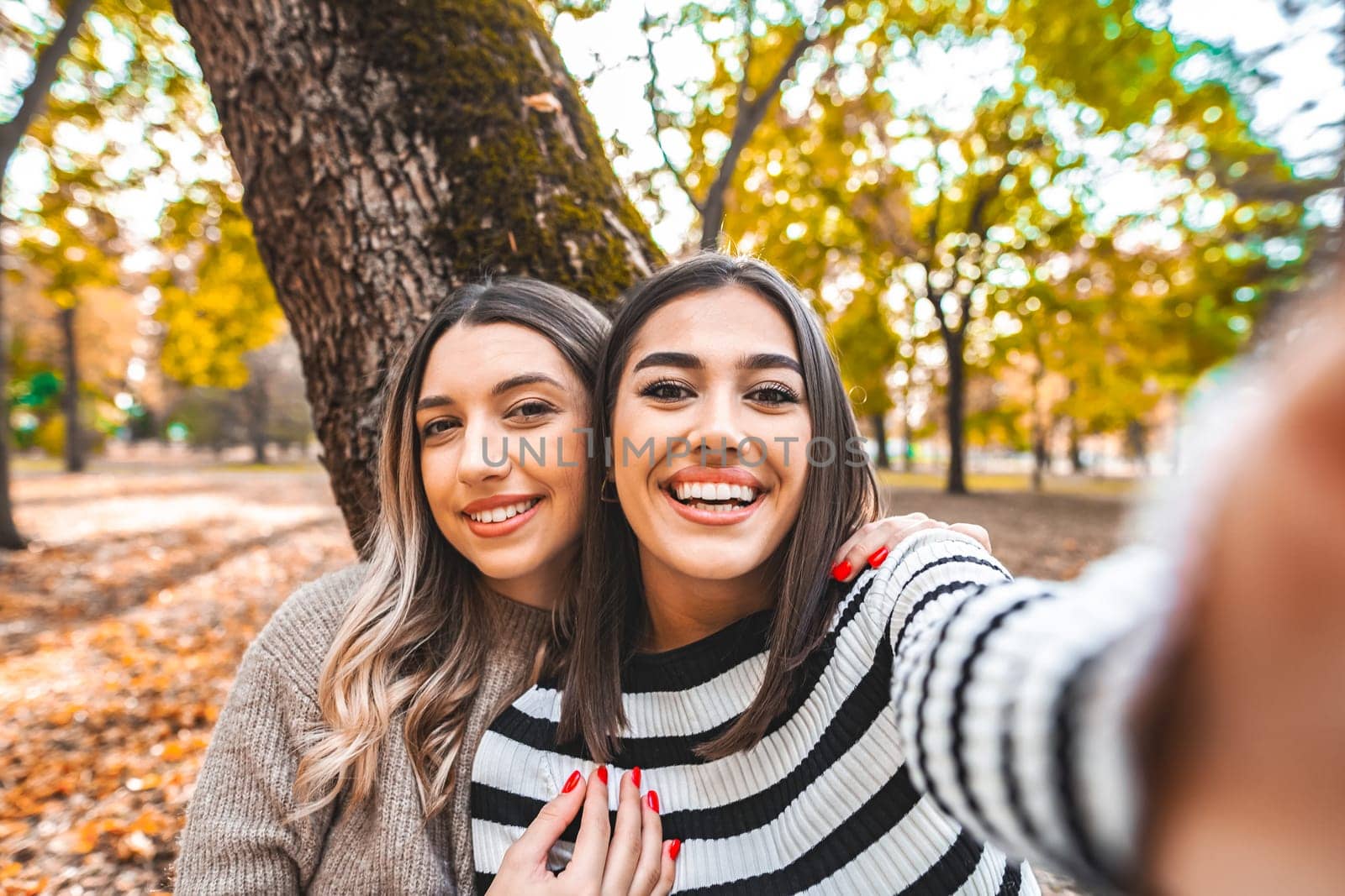 Two women, one brunette and one redhead, are smiling and posing while taking a selfie with a phone in a park. They are standing together in front of green trees under a blue sky.