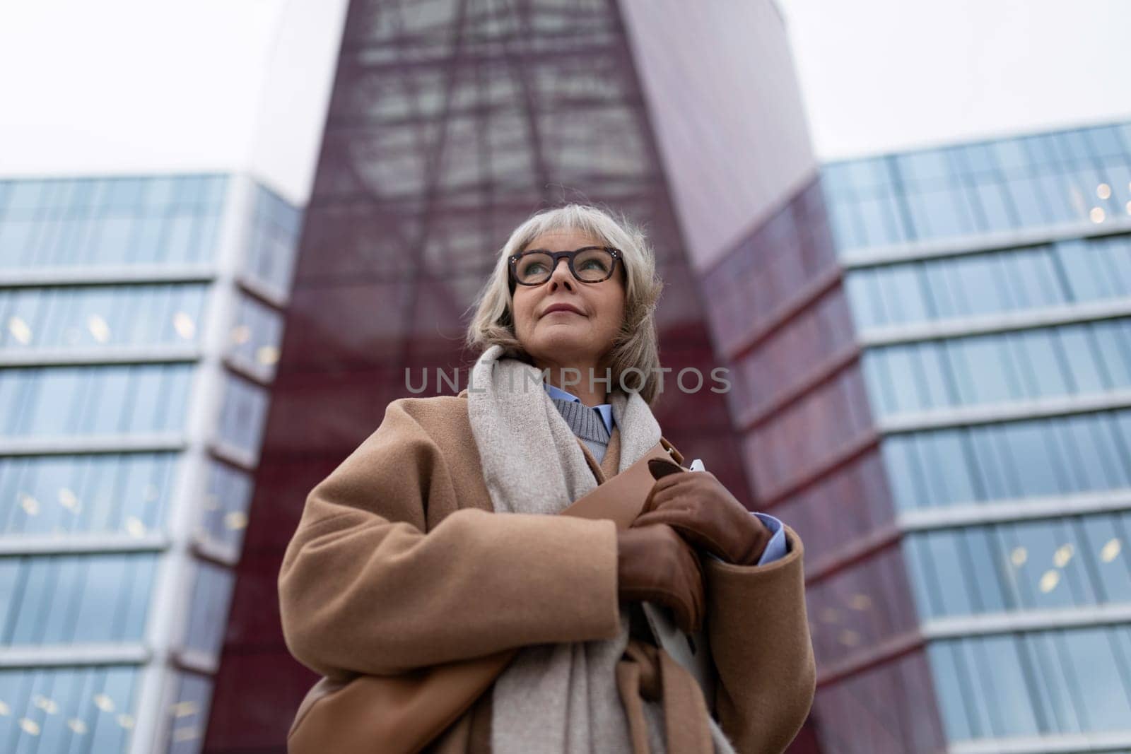 A stylish woman with gray hair and glasses poses gracefully in front of contemporary buildings, dressed warmly in a tan coat and scarf while showcasing urban elegance.