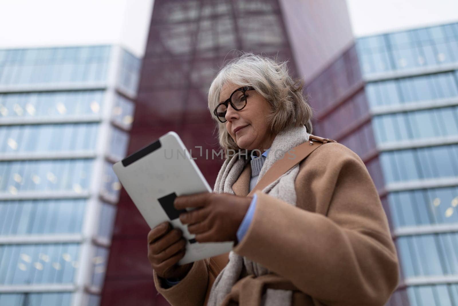 A mature woman with gray hair and glasses is focused on her tablet while standing in front of a contemporary glass structure. The scene captures an afternoon vibe.