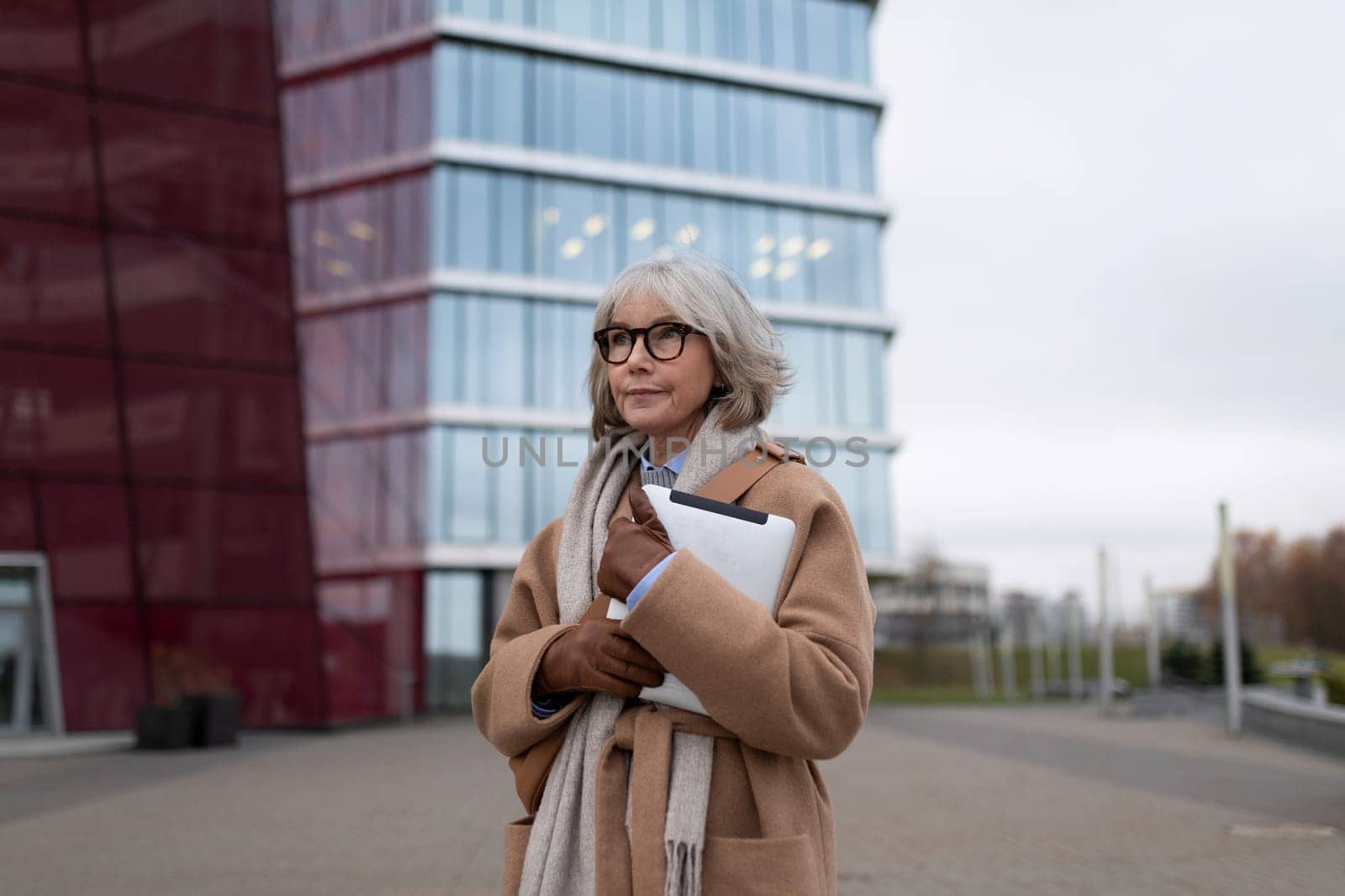 Dressed in a stylish coat and scarf, an older woman stands confidently in front of a contemporary glass building, preparing for her next business engagement on an overcast day.