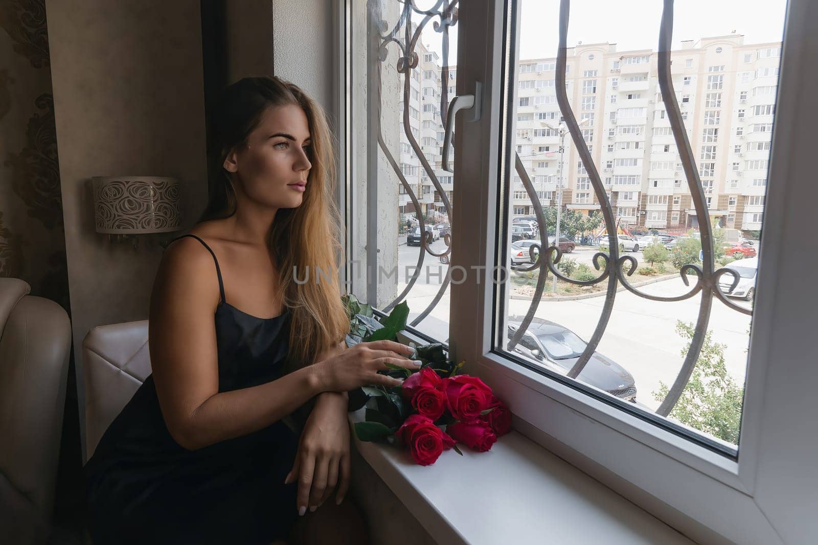A woman in a black dress is sitting in front of a window with a bouquet of red roses