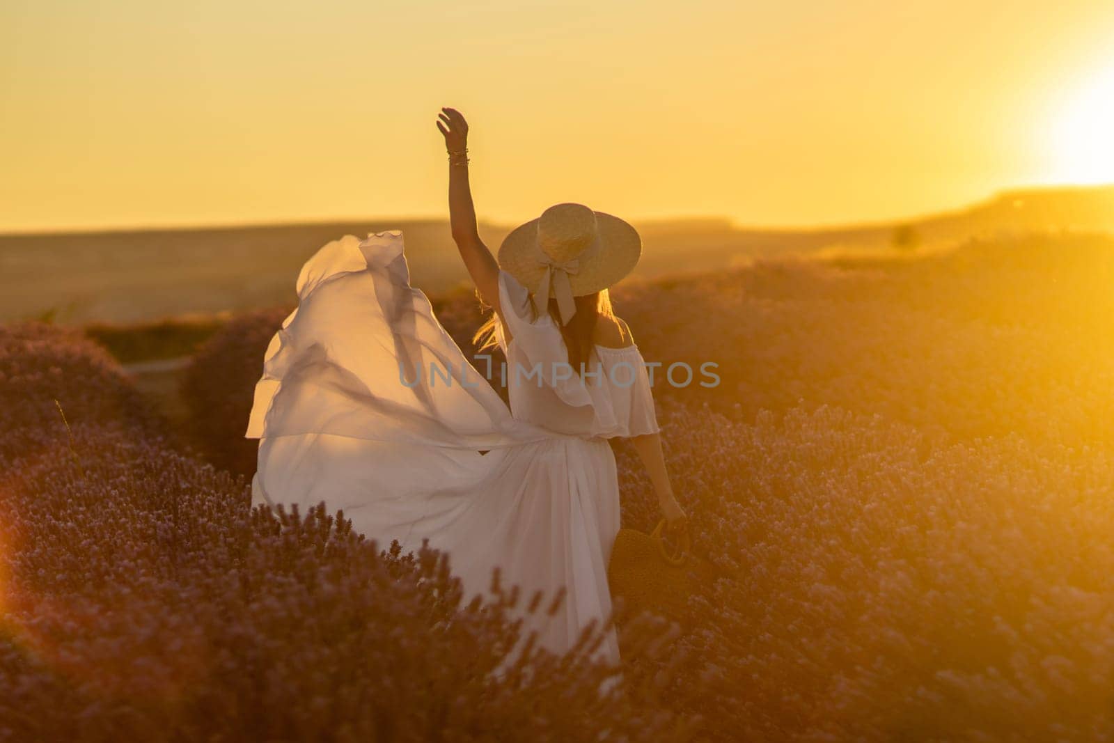 Here's a caption under 150 characters describing the image: Woman in white dress and straw hat joyfully dances in a lavender field at sunset. Golden light bathes the scene.